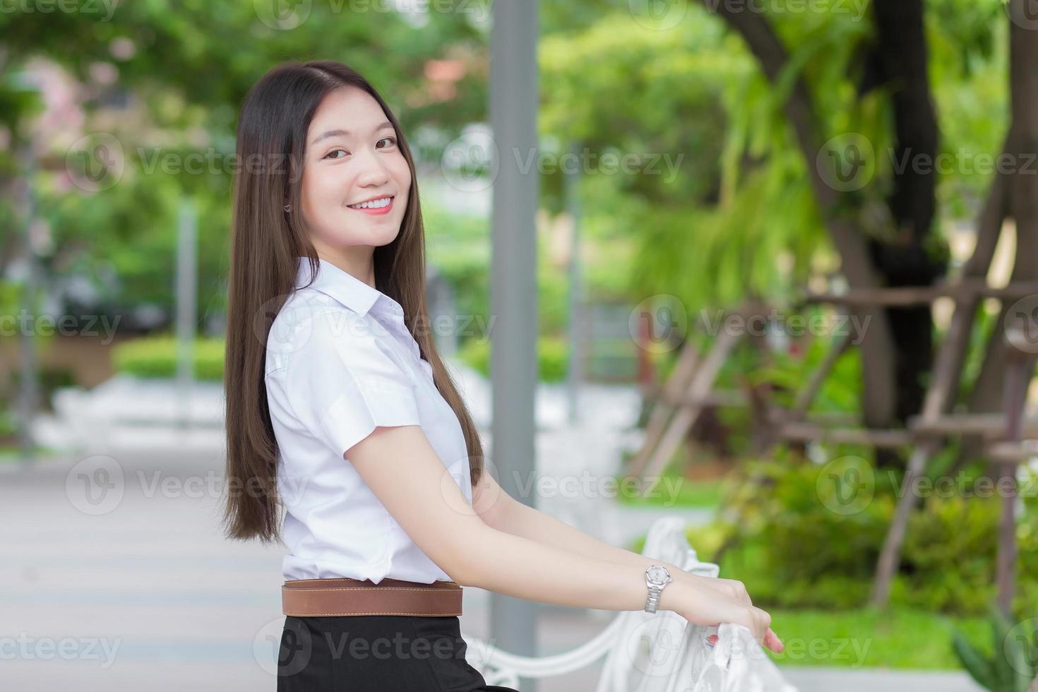 porträtt av vuxen thai student i universitetsstudentuniform. asiatisk vacker flicka sitter och ler glatt på utomhusuniversitet med en bakgrund av trädgårdsträd. foto