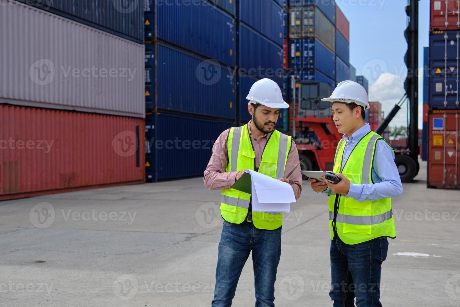 två professionella asiatiska manliga arbetare i säkerhetsuniformer och hjälm arbetar på en logistikterminal med många travar av containrar och lastar kontrollerar fraktgods för godstransportindustrin. foto