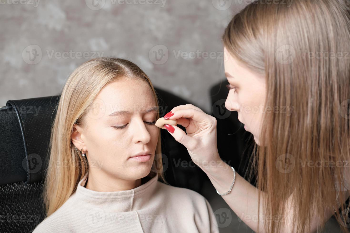 kvinna make up artist använder svamp för att applicera foundation eller bb cream på kvinnans ansikte. hudtonskorrigering, ansiktssminkning i skönhetsstudio foto