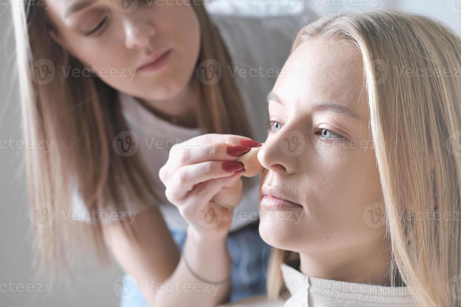 visagiste eller make-up artist som använder svamp för att applicera cc cream på ung kvinnas ansikte. blond vacker kvinna i skönhetsstudio foto