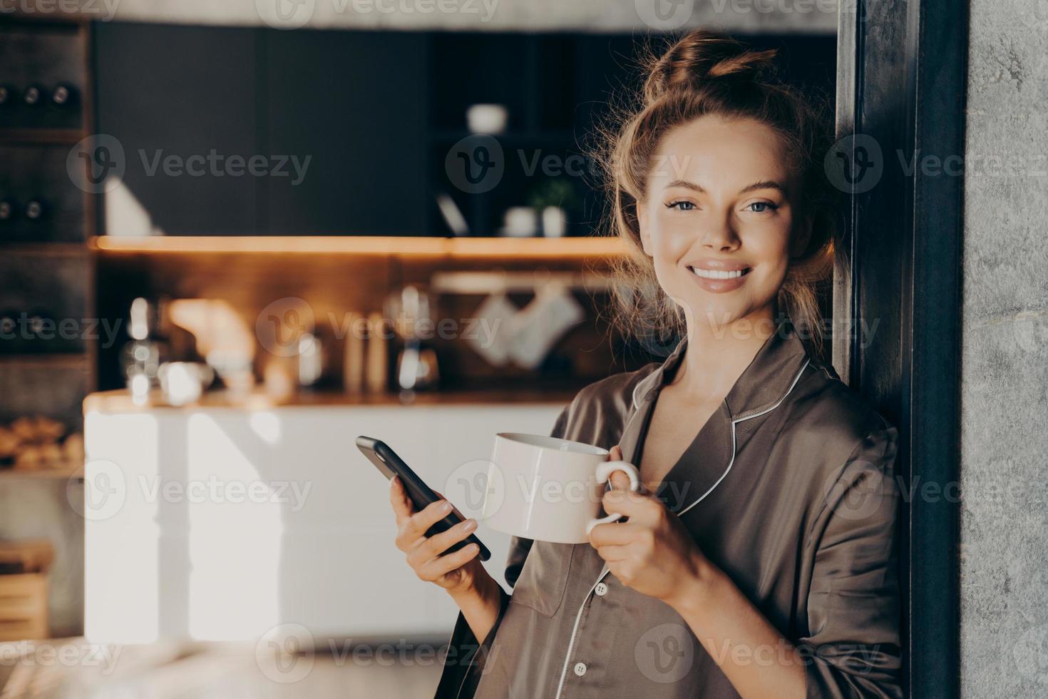 härlig glad ung kvinna som har sitt morgonkaffe medan hon kollar nya e-postmeddelanden och aviseringar på smartphone foto