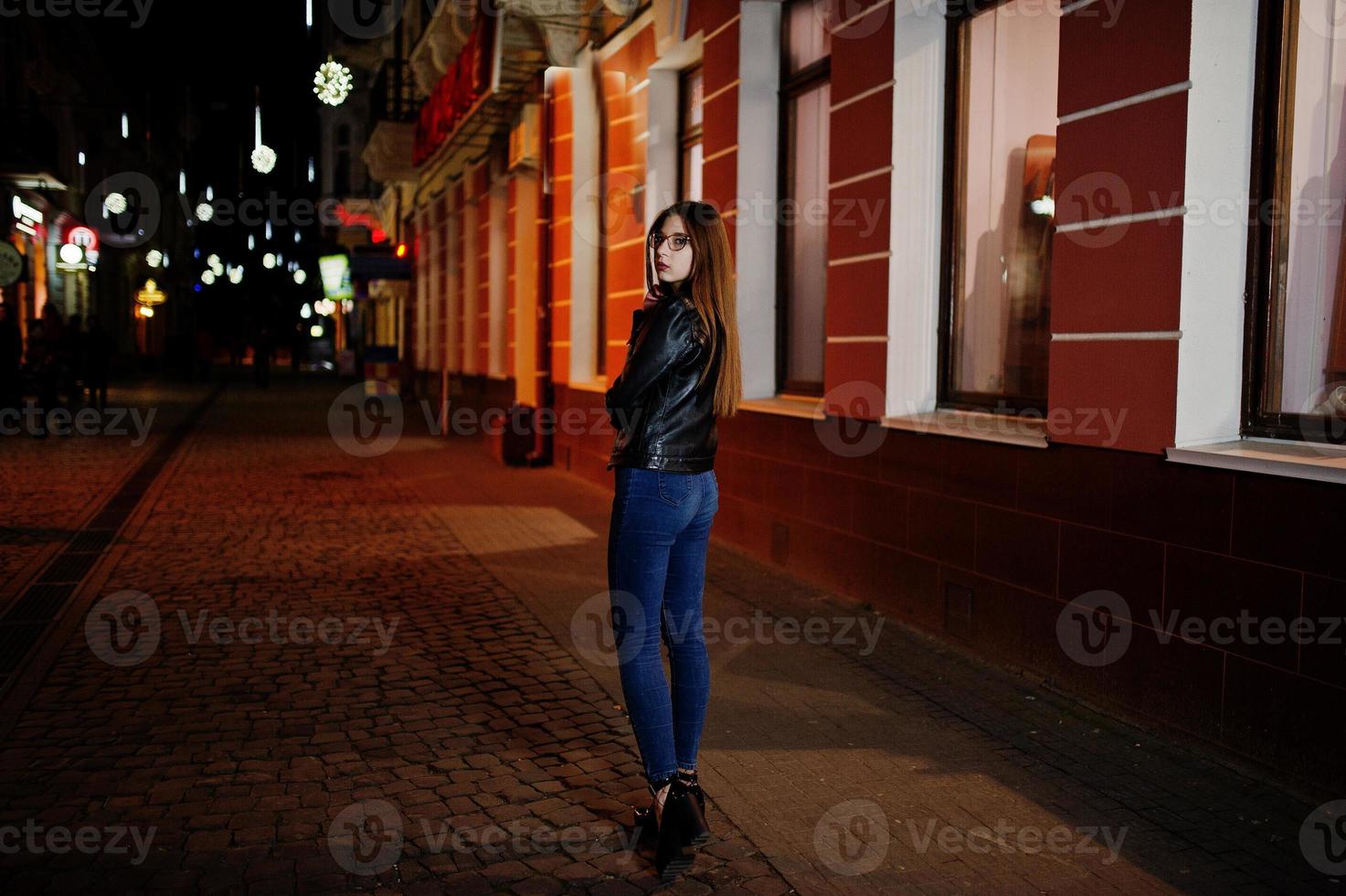 nattporträtt av flickmodell slitage på glasögon, jeans och skinnjacka, med ryggsäck, mot stadens gator. foto