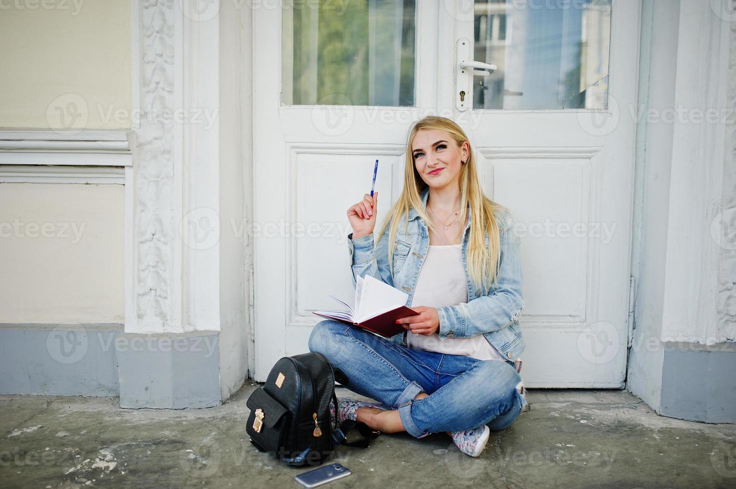 blond flicka bära jeans med ryggsäck poserade mot gamla dörren med dagbok och skriva något. foto