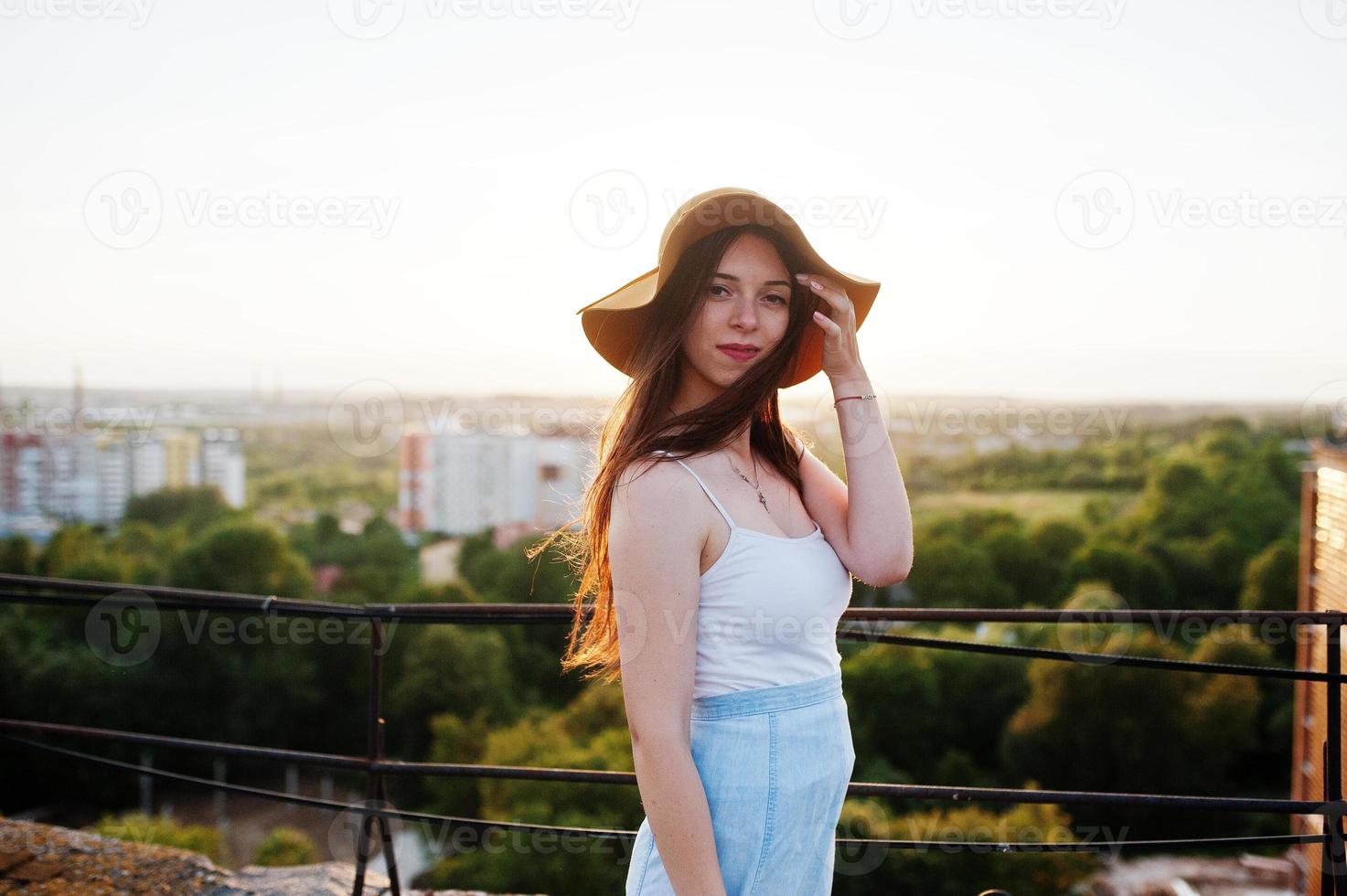 porträtt av en vacker ung kvinna i vit t-shirt och blå kjol poserar på taket med sin orange hatt vid solnedgången. foto