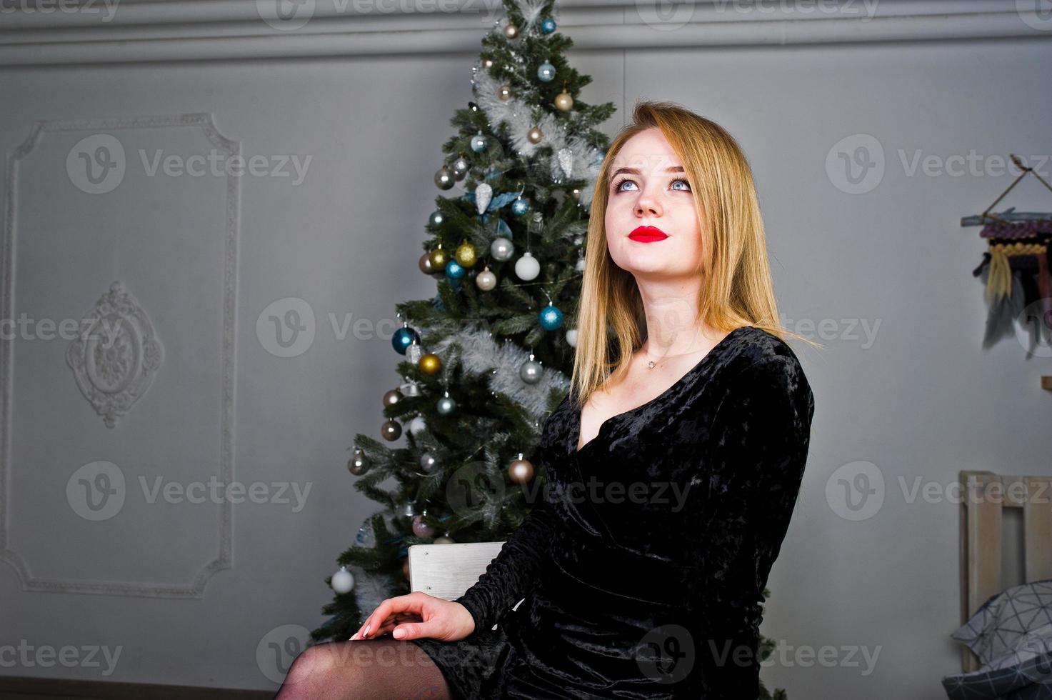 elegant blond flicka bär på svart klänning mot nyårsträd med juldekoration. foto