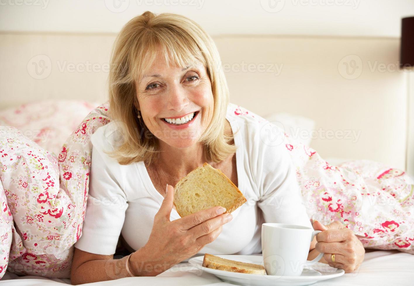äldre kvinna snugglade under täcken och äter frukost foto