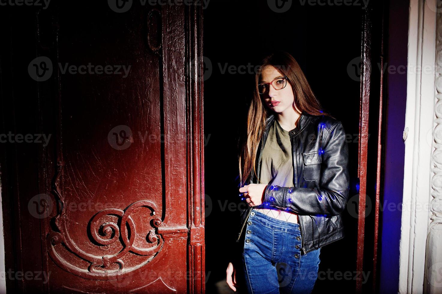 nattporträtt av flickmodell slitage på glasögon, jeans och skinnjacka, med blå krans på henne. foto