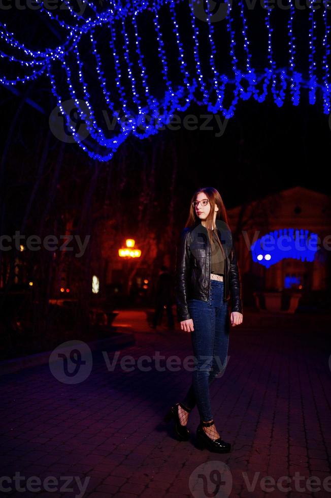 nattporträtt av flicka modell slitage på glasögon, jeans och skinnjacka mot blått ljus krans av stadsgatan. foto