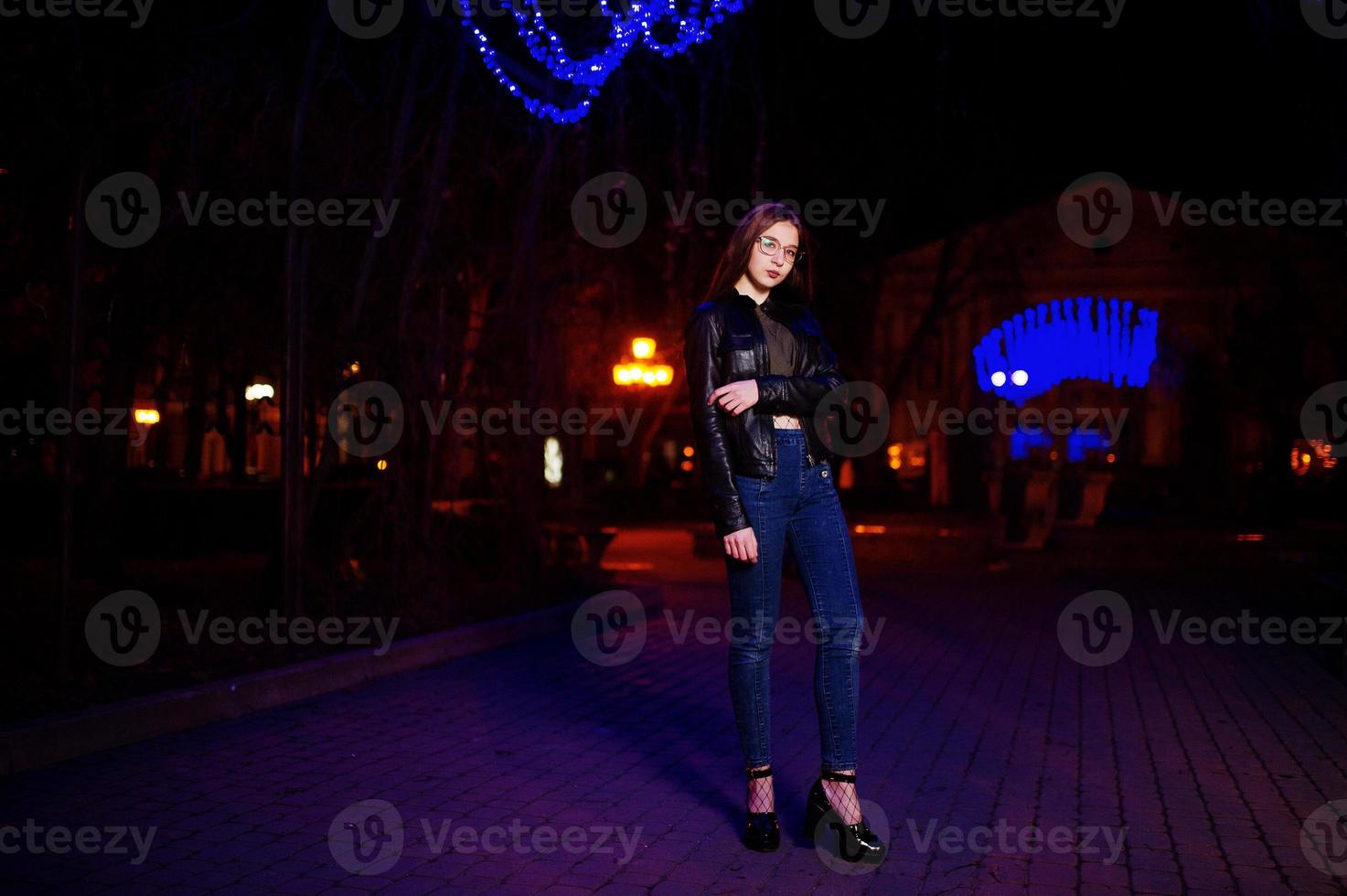 nattporträtt av flicka modell slitage på glasögon, jeans och skinnjacka mot blått ljus krans av stadsgatan. foto
