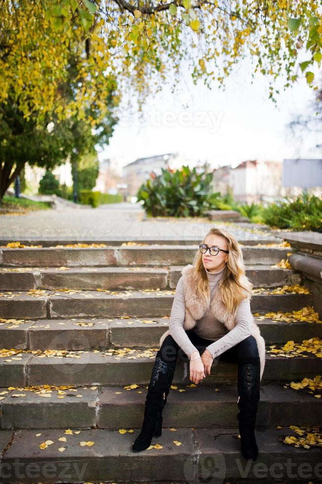 blond flicka på päls och glasögon sitter på trappan med gula löv. foto