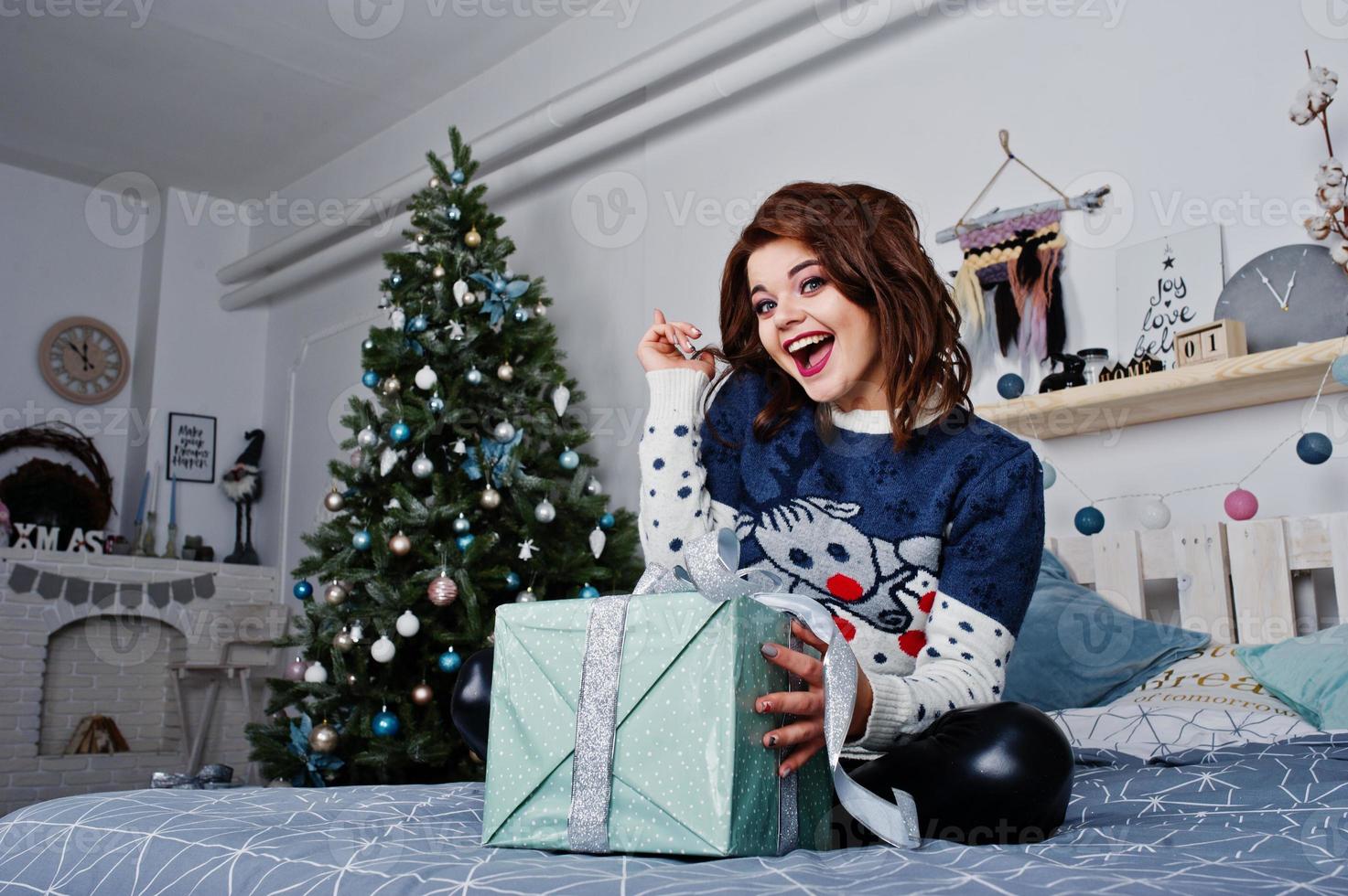 flicka bär varm tröja sitter på sängen mot nyår träd med presenter ruta till hands på studio. glad vintersemester koncept. foto
