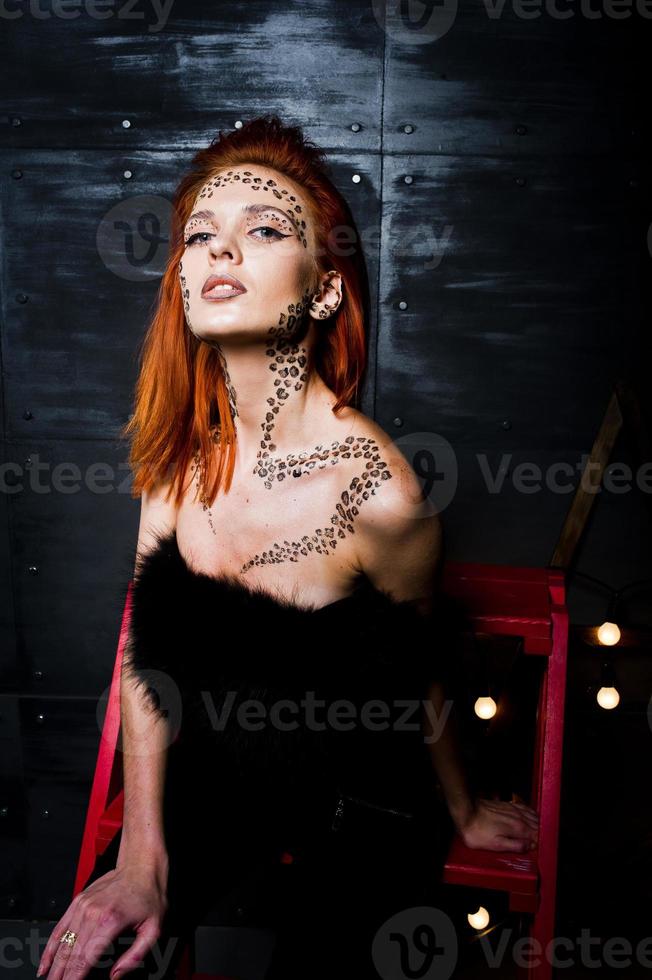 modell rödhårig tjej med ursprungligen smink som leopardrovdjur mot stålvägg. studio porträtt på stege. foto