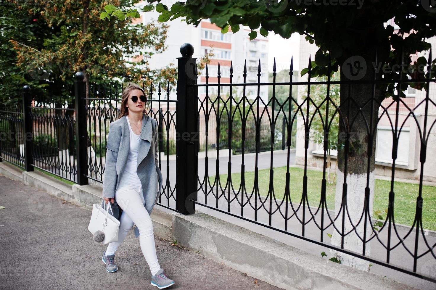 flicka i grå rock med solglasögon och handväska går på gatan mot järn staket. foto