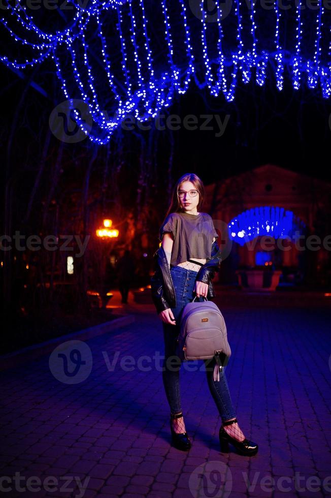 nattporträtt av flicka modell slitage på glasögon, jeans och skinnjacka, med ryggsäck i händerna, mot blått ljus krans av stadsgatan. foto