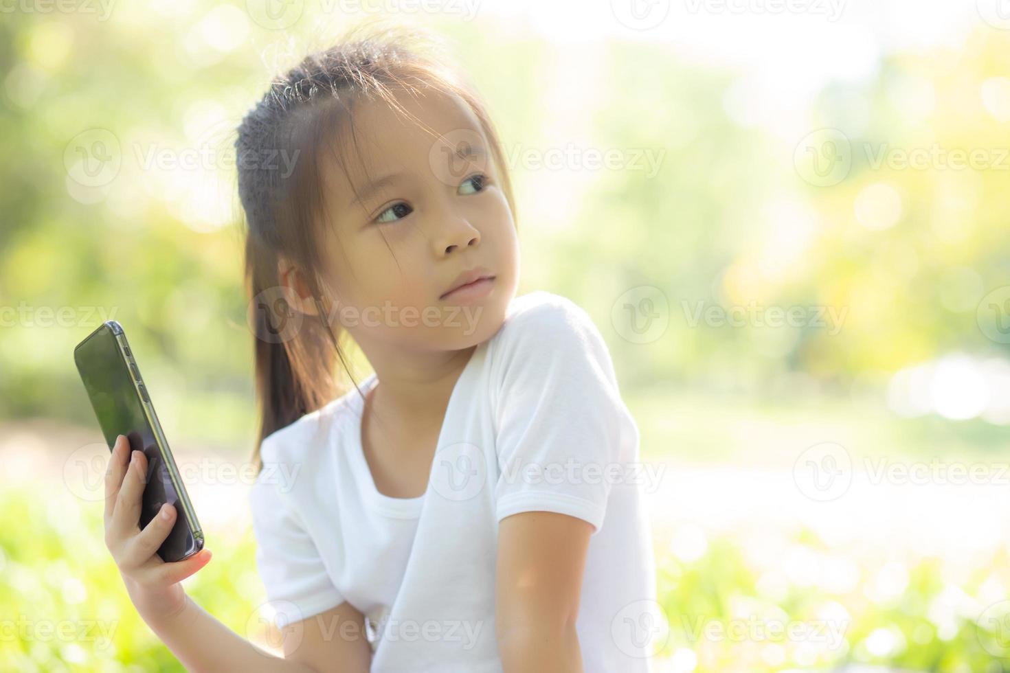 vackert asiatiskt barn som ler med smart mobiltelefon i trädgården, barn har passion för att spela digitalt spel på smartphone på internetnätverk i parken med glad, livsstil och teknikkoncept. foto