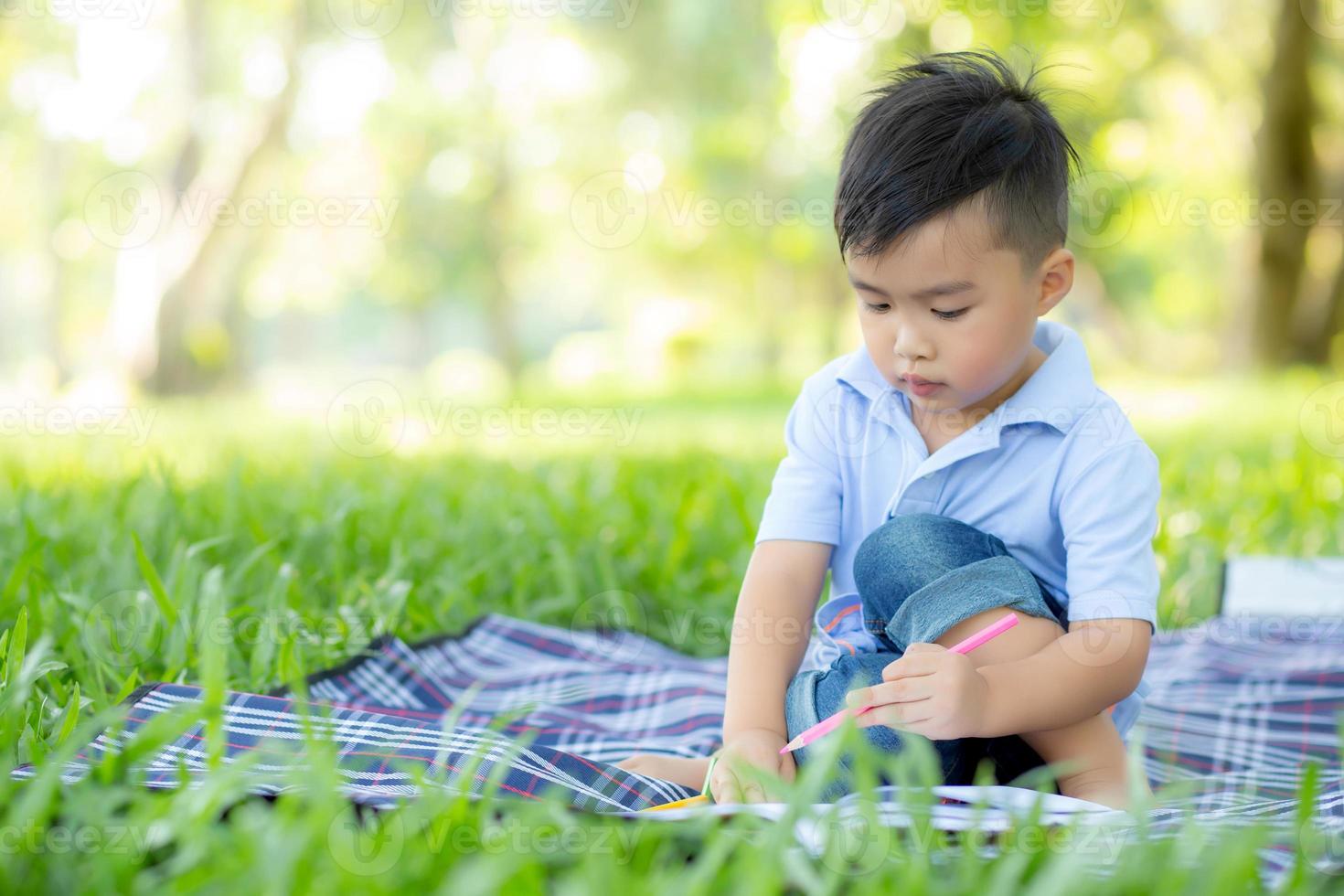 ung liten asiatisk pojke som läser och skriver bok i parken, asia kid läxor och studera på sommaren, barn koppla av med att rita på anteckningsboken i semester, utbildning och utveckling koncept. foto