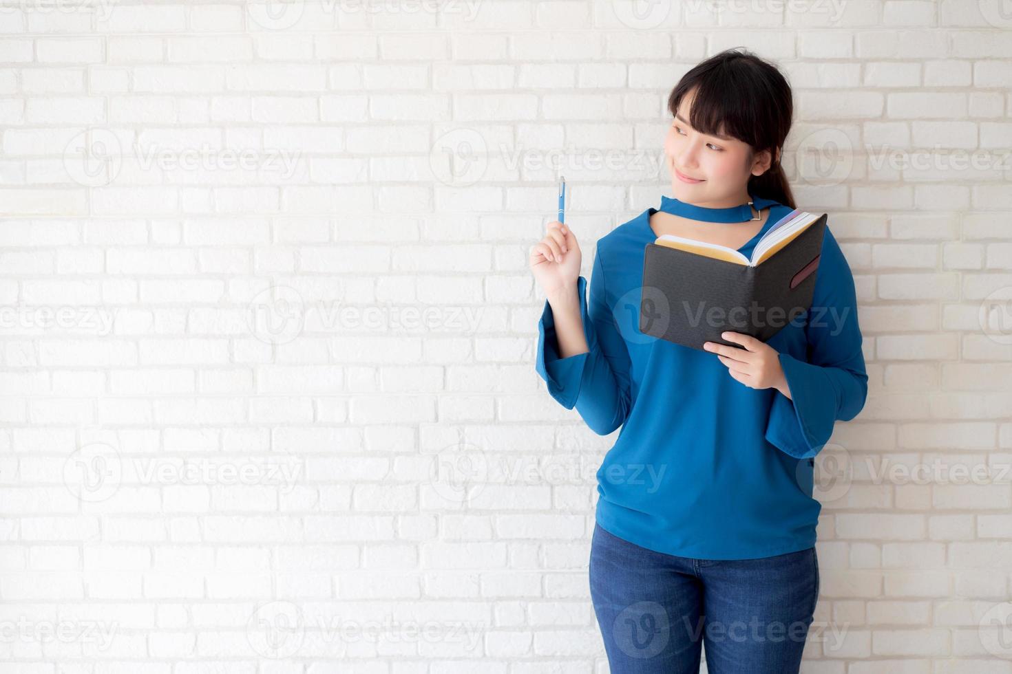 vacker asiatisk kvinna som ler står och tänker och skriver anteckningsbok på betongcement vit bakgrund hemma, flickläxa om bok, utbildning och livsstilskoncept. foto