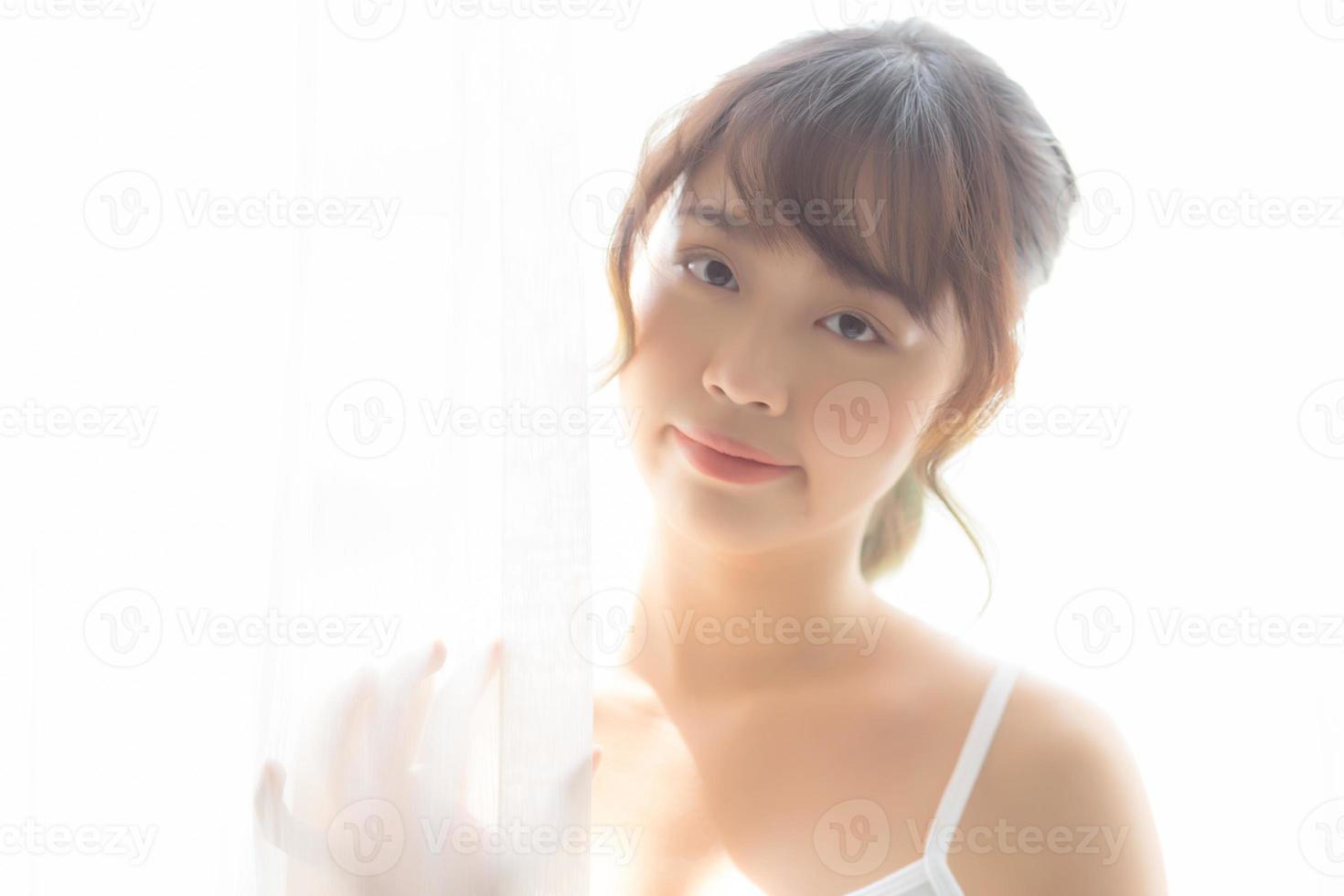 porträtt av vacker ung asiatisk kvinna som står i fönstret och ler medan hon vaknar med soluppgången på morgonen, flicka glad med friskhet och glad, livsstil och slappna av koncept. foto