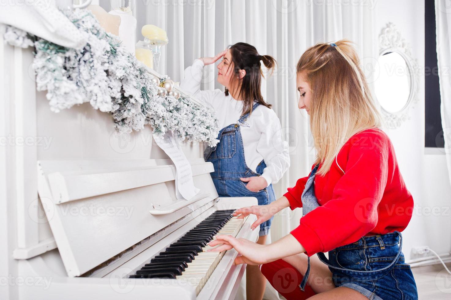 två roliga vackra tjejer vänner bär i overall jeans shorts och damasker mot piano med nyårsdekoration. foto