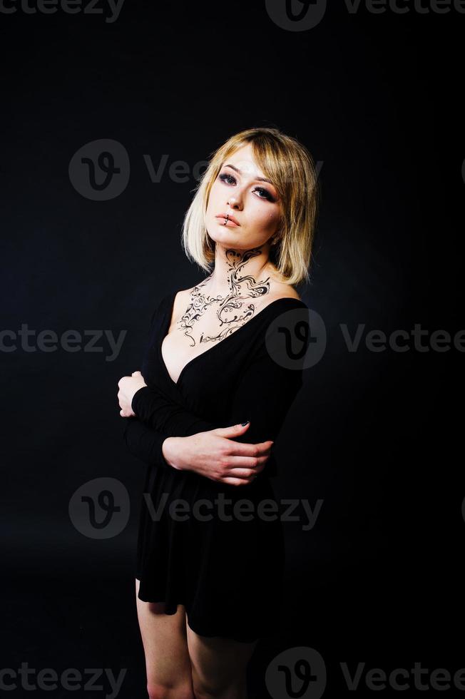 studio porträtt av blond tjej med ursprungligen smink på halsen, bär på svart klänning på mörk bakgrund. foto
