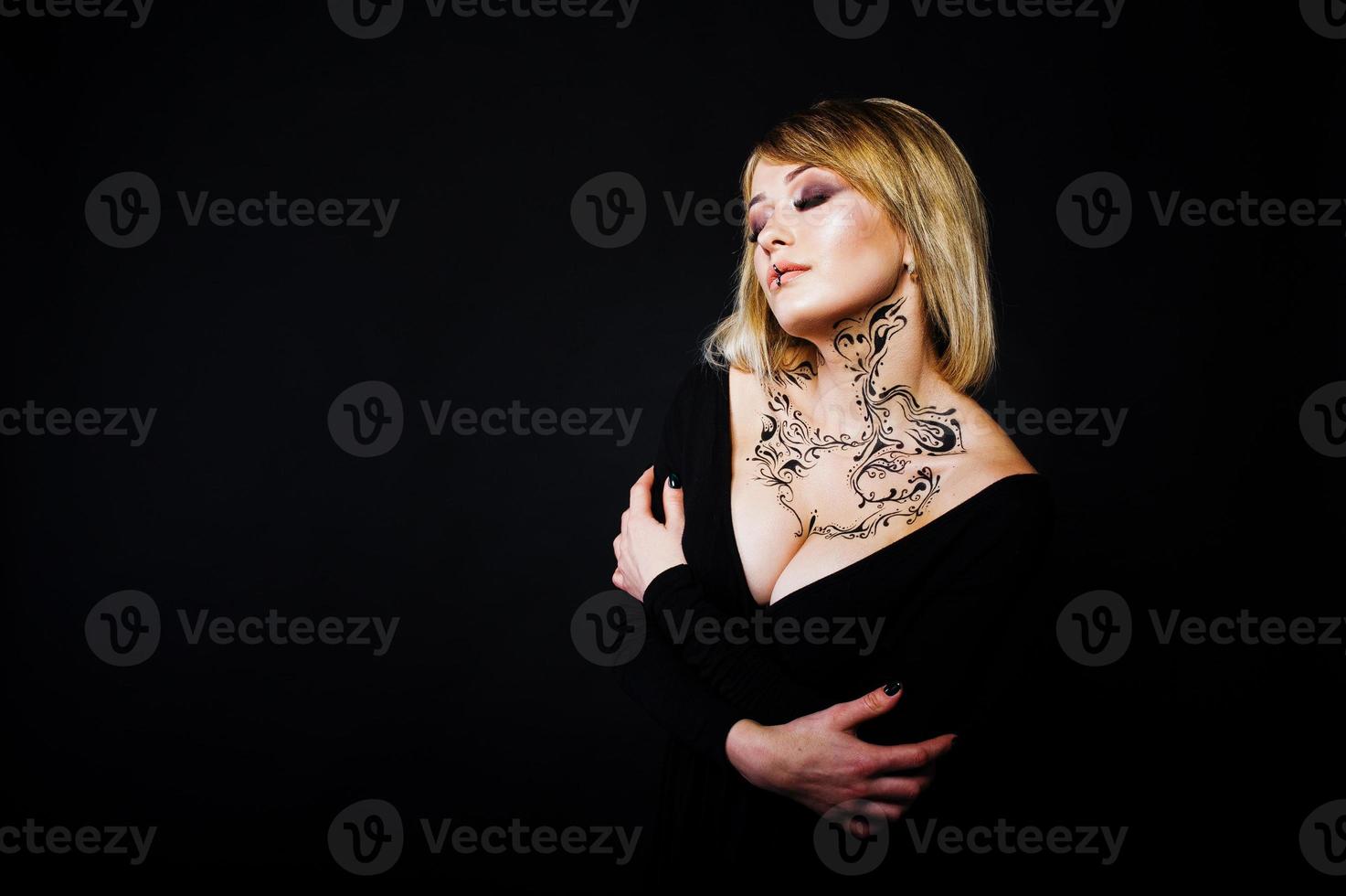 studio porträtt av blond tjej med ursprungligen smink på halsen, bär på svart klänning på mörk bakgrund. foto