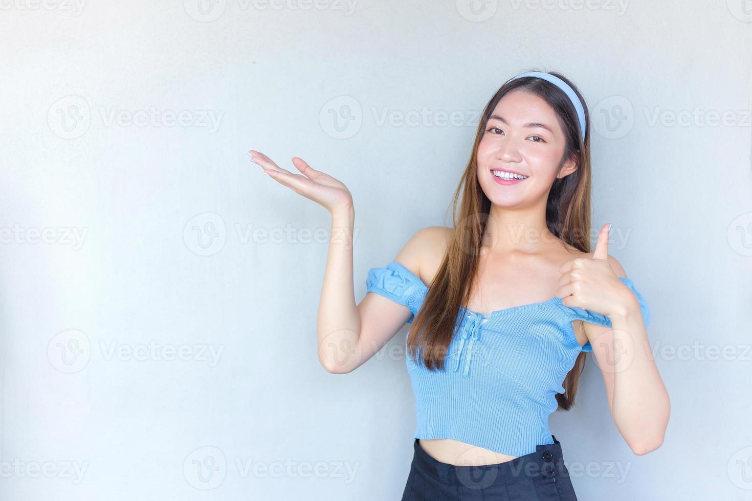 ung vacker asiatisk kvinna i en blå skjorta agerar tummen upp som en bra symbol och en annan hand visar sig presentera något på vit bakgrund. foto