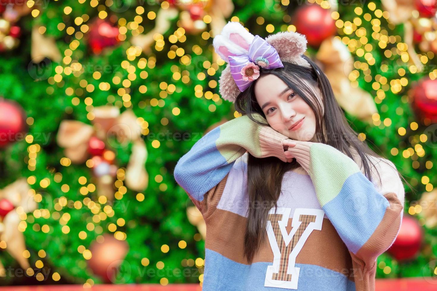 en vacker asiatisk kvinna i en färgglad tröja står leende och glad framför julgranen med bokeh som bakgrund i temat för att fira jul och gott nytt år foto