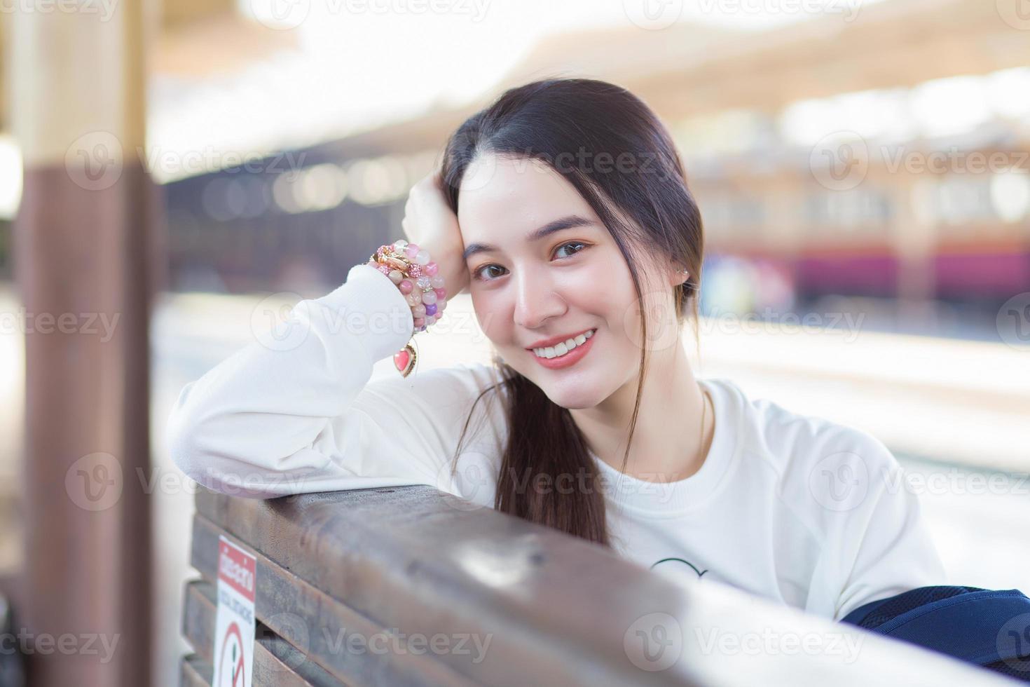 asiatisk vacker kvinna i en långärmad vit skjorta och en hatt sitter glad smilie på tågstationen och väntar på att tåget ska komma. foto