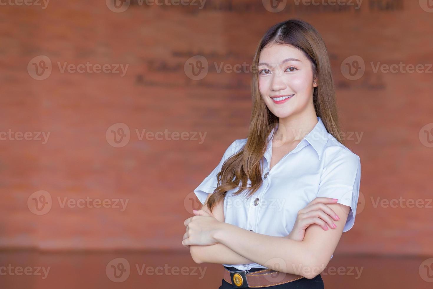 porträtt av vuxen thai student i universitetsstudentuniform. asiatisk vacker flicka står leende med armarna i kors på en tegel bakgrund. foto