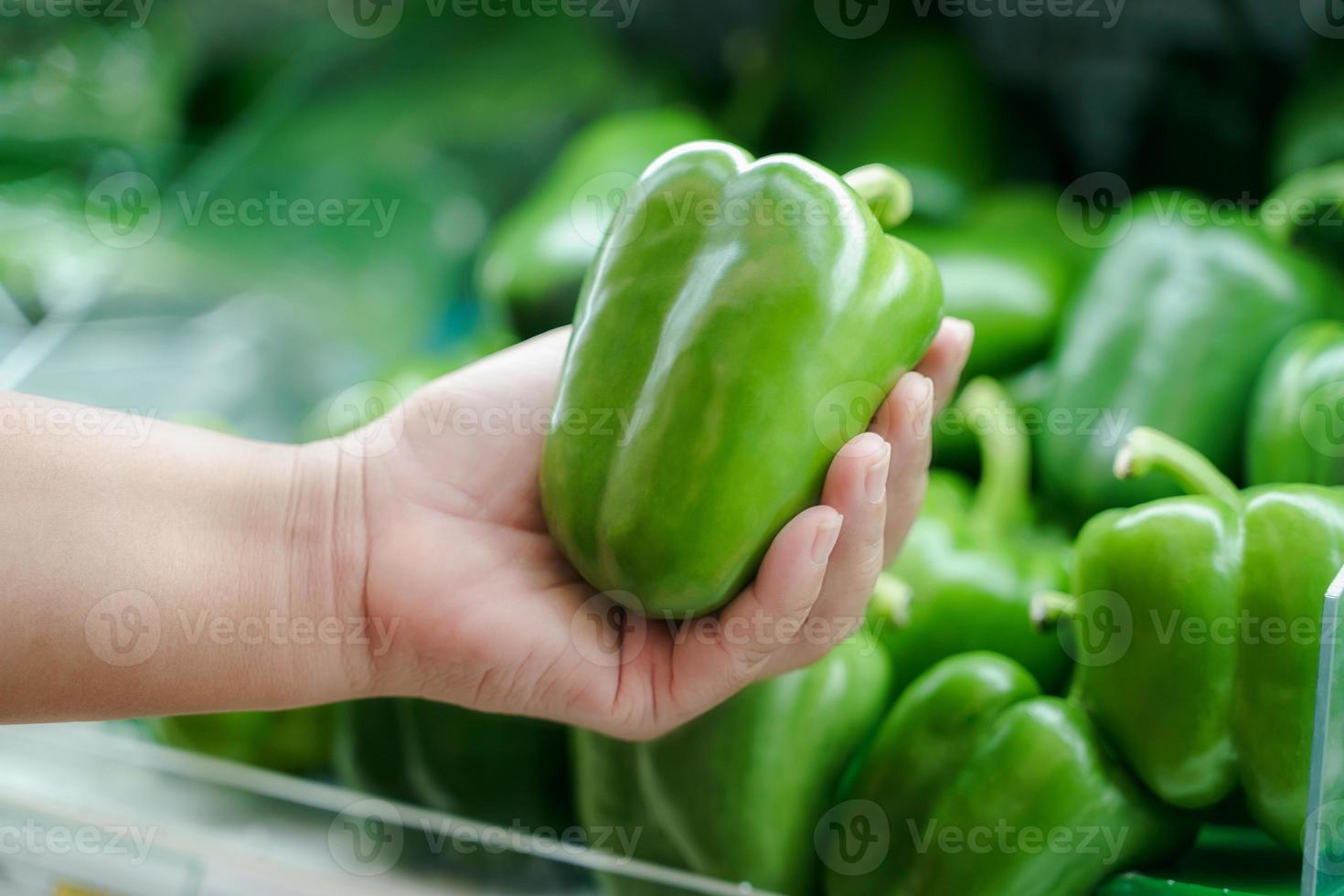 närbild kvinna hand håller färsk grön paprika på hyllan på snabbköpet. foto