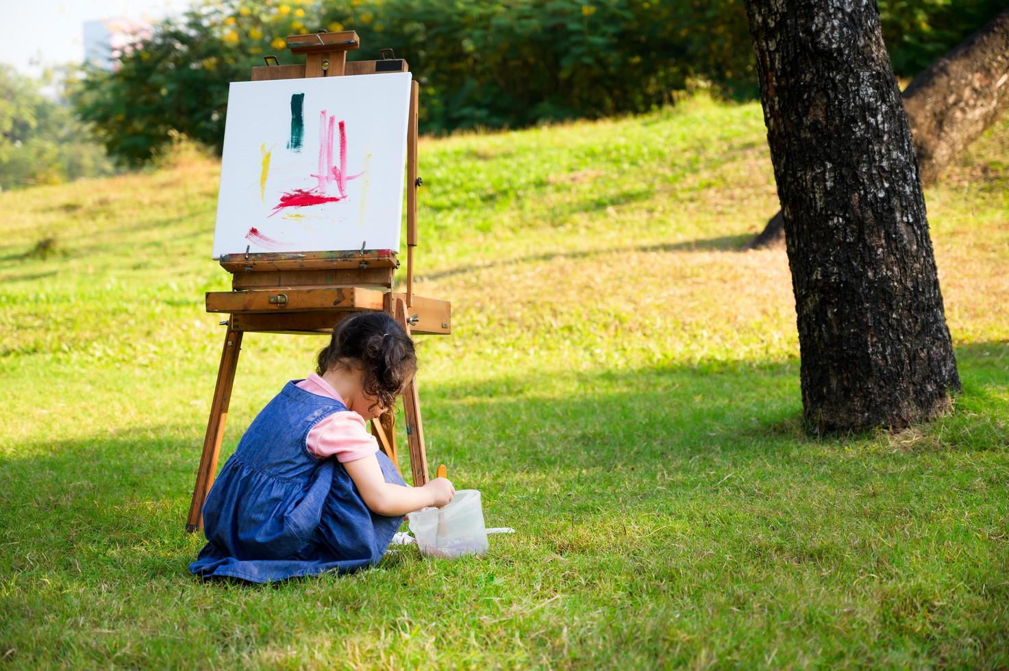 en liten flicka sitter på gräset och målad på duken placerad på ett ritställ foto