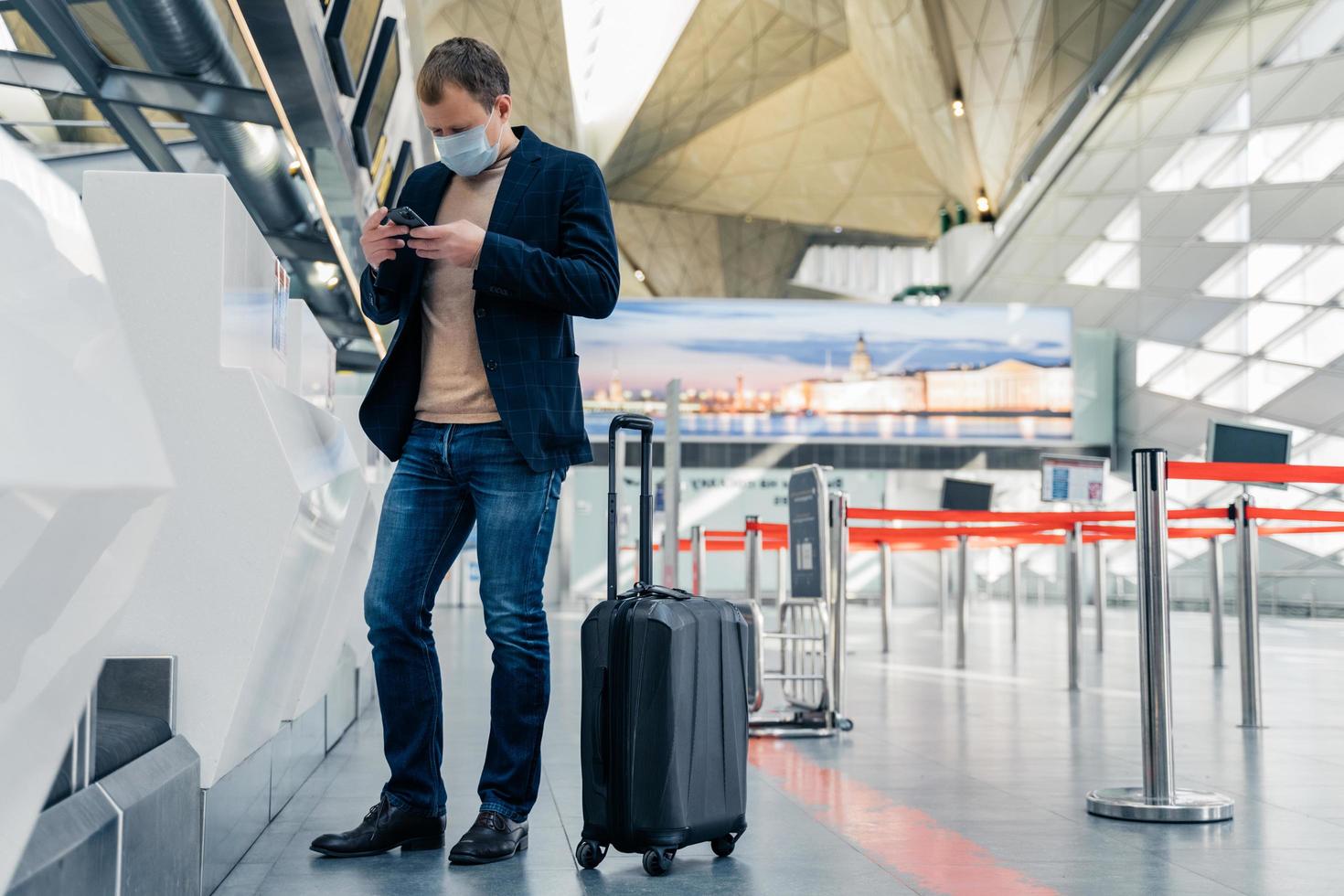 fullängdsbild av man passagerare står nära resväska på hjul på flygplats läser nyheter online på mobil bär skyddande medicinsk mask får reda på stängning av landsgränser på grund av virus foto