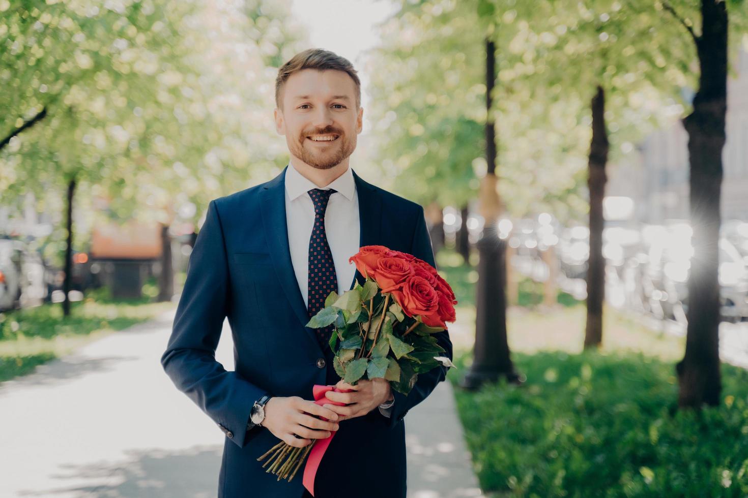 glad ung man i elegant kostym med bukett blommor som går i parken foto