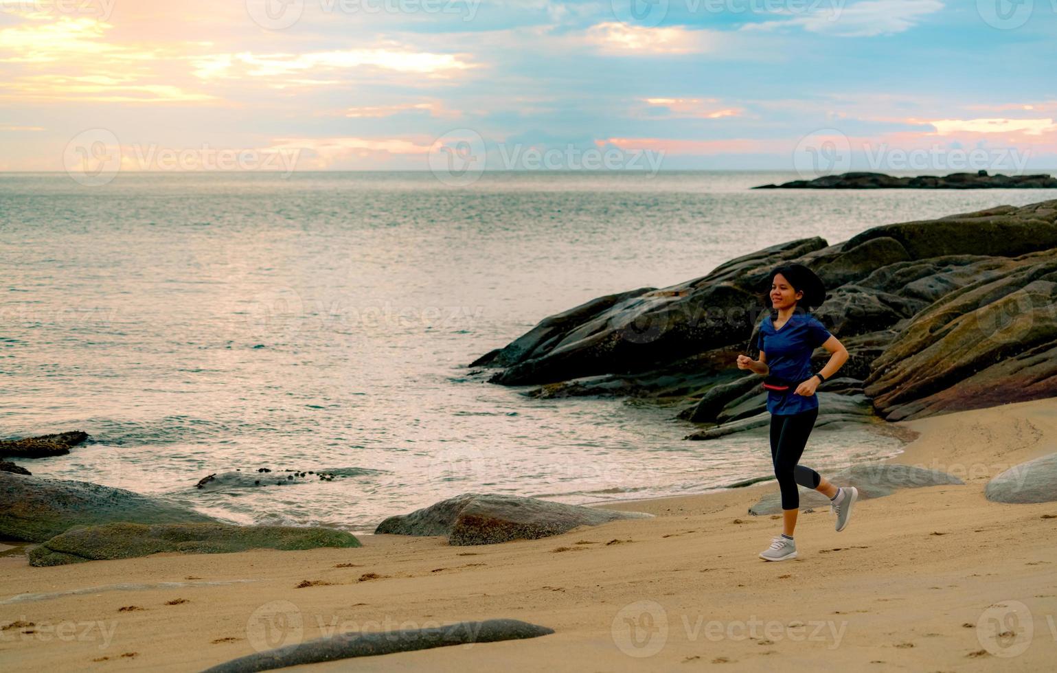 asiatisk kvinna kör på tropiska havet stranden på morgonen med vacker soluppgång himmel. passa frisk kvinna utomhus träning. fit tjej bär smart band och midjeväska. kvinna kör för konditionsträning. foto