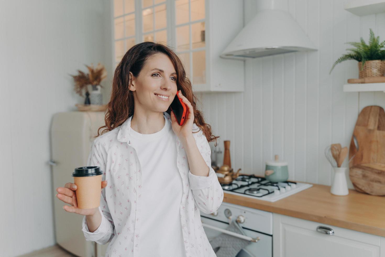 ung kaukasisk kvinna med telefon har kaffe och samtal i sitt eleganta kök hemma. foto