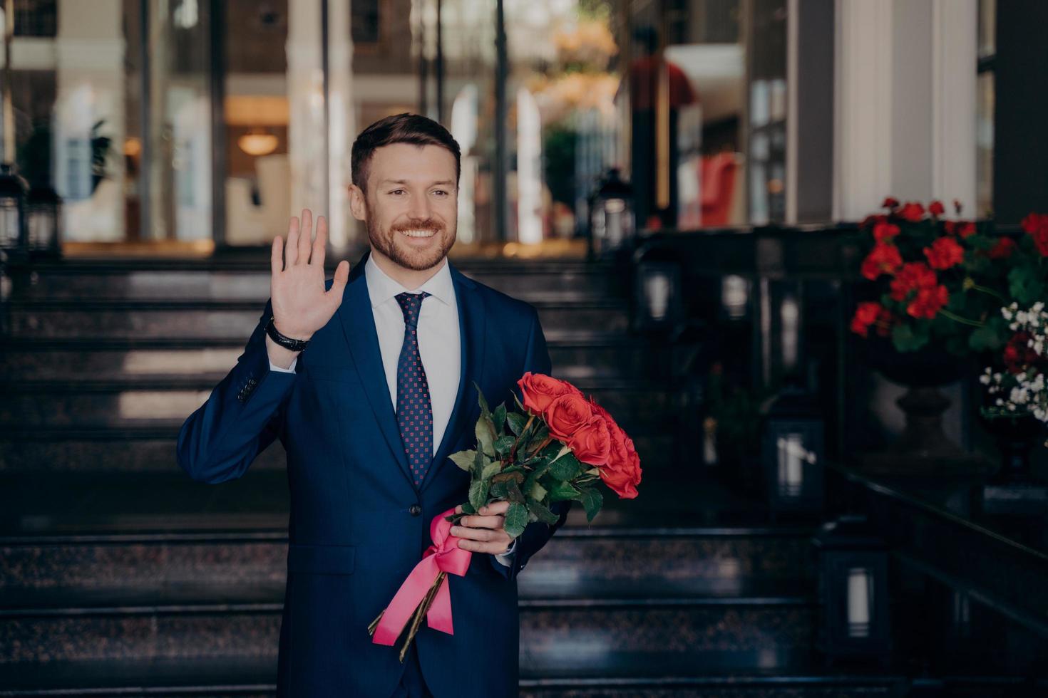 attraktiv man väntar på gatan i elegant mörkblå kostym med röd ros blombukett foto