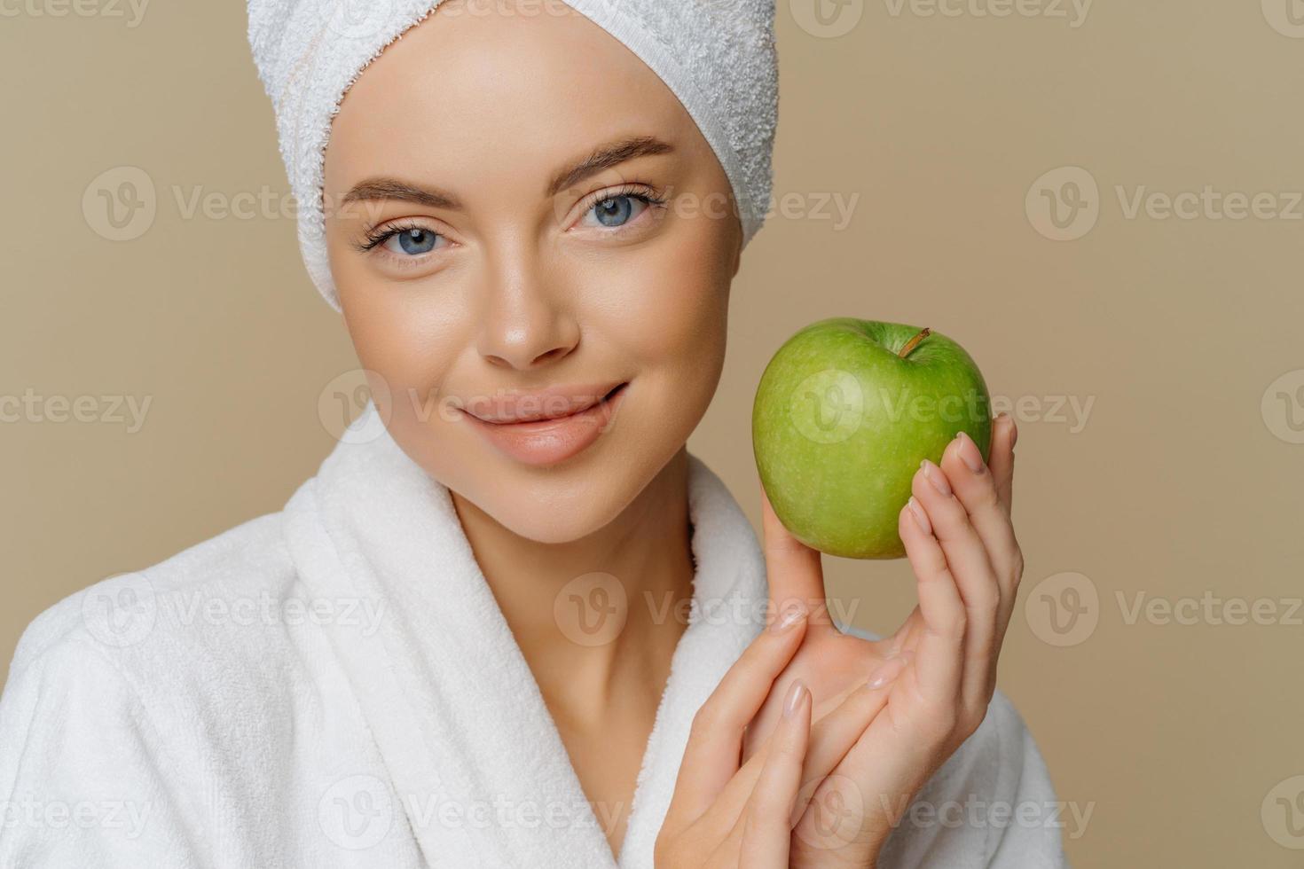 närbild av blåögd kvinna med minimal makeup välskött hy fräsch ren hud klädd i vit rock har grönt äpple isolerat över brun bakgrund. naturlig kosmetika koncept. foto