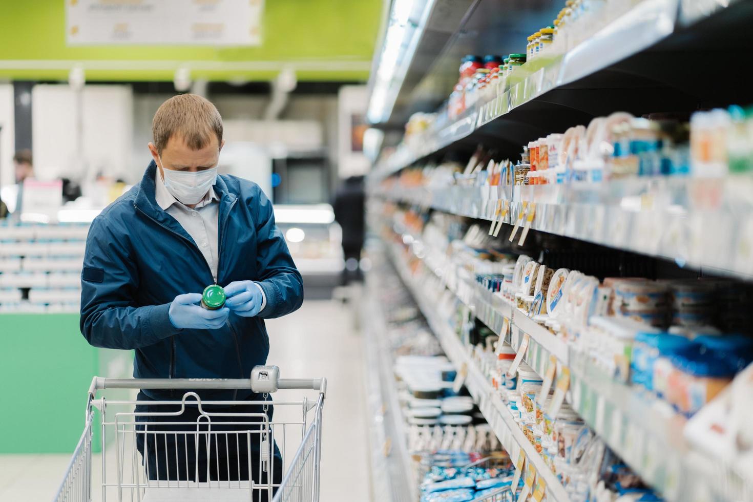 horisontell bild av vuxen man bär skyddsmask, läser etiketten på produkten, handlar under coronavirusutbrottet, köper nödvändig mat i lokal butik. människor, virus, sjukdom, köpkoncept foto