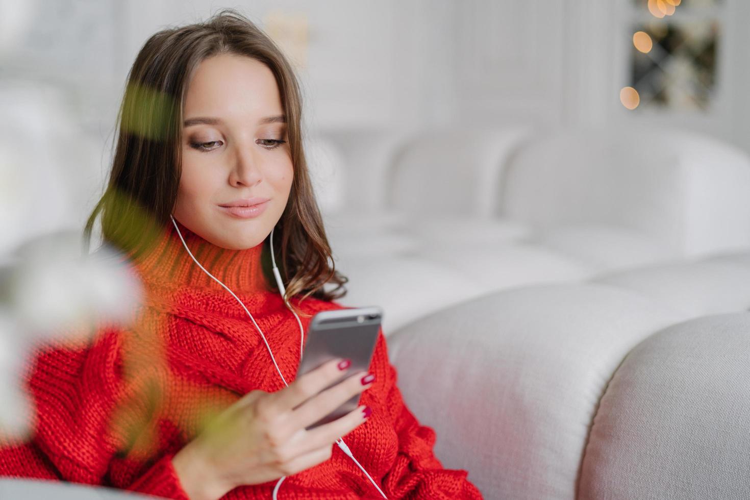 vacker ung kvinna håller mobiltelefonen, lyssnar på musik från spellistan, surfar på sociala nätverk och meddelanden med vänner, njuter av en lugn atmosfär hemma, sitter i en bekväm soffa. livsstilskoncept foto