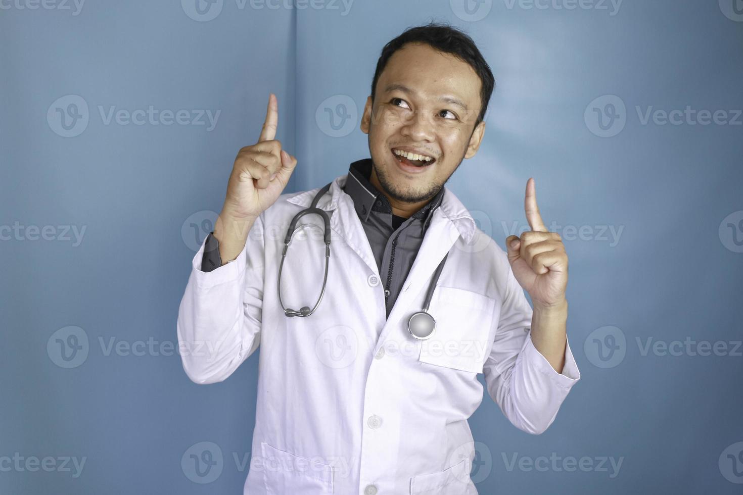 glad ung asiatisk man doktor, en läkare ler och pekar uppåt på ett kopia utrymme isolerat över blå bakgrund foto