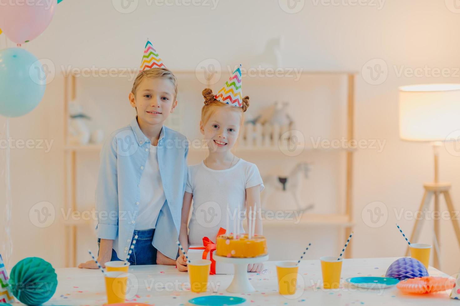 glad pojke och hans lilla ingefära syster iklädda festkläder, festhattar, firar födelsedag tillsammans, omgivna av tårta, present och koppar dryck, har gott humör under speciella tillfällen foto