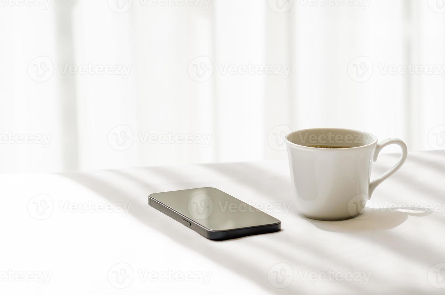 selektiv inriktning av en kopp svart kaffe och smartphone sätta på vitt lakan med skuggan av gardin på morgonen. avkopplande hemma koncept. foto