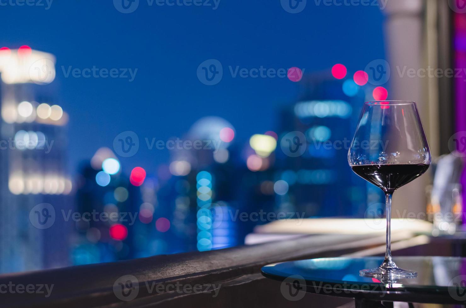 ett glas rött vin på bordet i takbaren med färgglad bokeh av stadsljus. foto