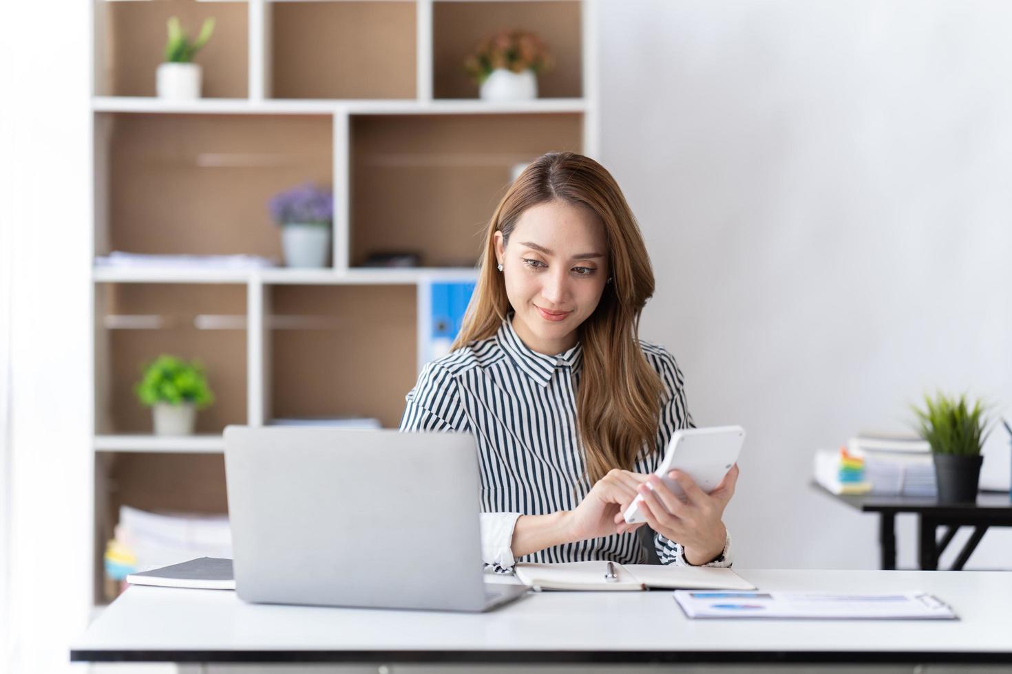 porträtt av asiatisk affärskvinna som betalar räkningar online med laptop på kontoret. vacker flicka med dator och checkhäfte, glad att betala räkningar. nystartade företag finansiella beräkna konto koncept foto