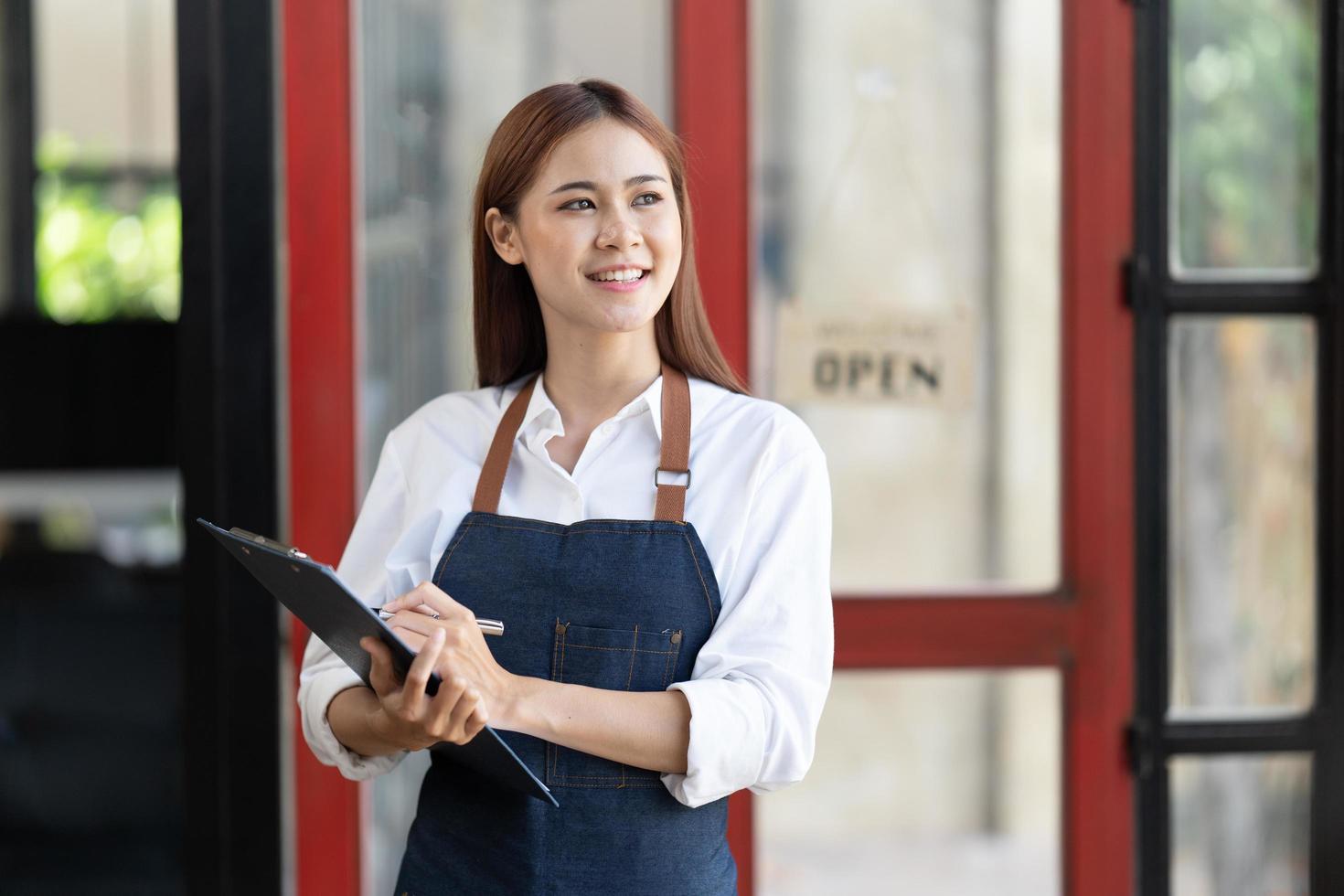 asiatisk glad affärskvinna är en servitris i ett förkläde, ägaren till kaféet står vid dörren med en skylt öppen och väntar på kunder. små affärsidé, kaféer och restauranger. foto