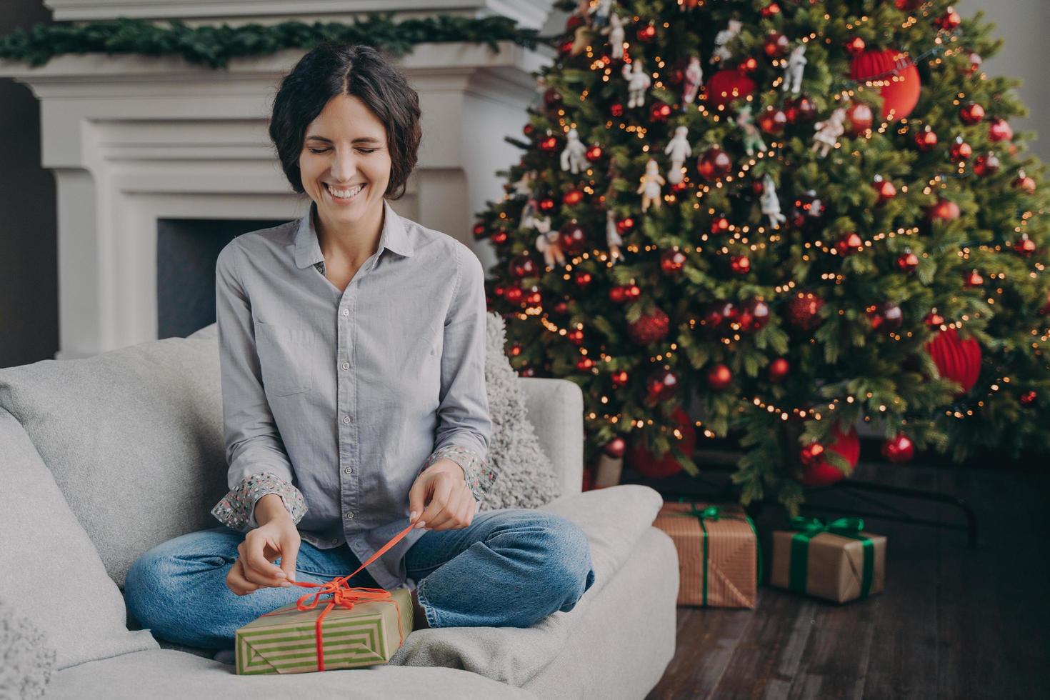 upprymd italiensk kvinna med slutna ögon som försöker packa upp julklappen när hon sitter på soffan hemma foto