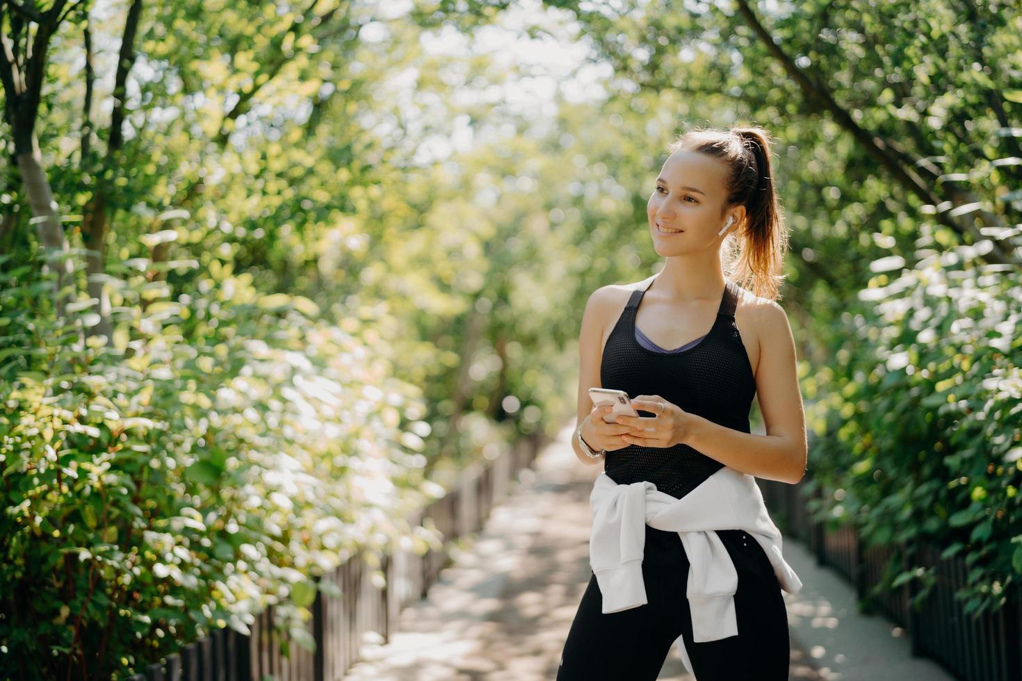 horisontell bild av aktiv sportig kvinna använder smartphone för att kontrollera resultat efter jogging klädd i aktivt slitage njuter av en varm solig dag lyssnar på musik via hörlurar. hälsosam livsstil koncept. foto