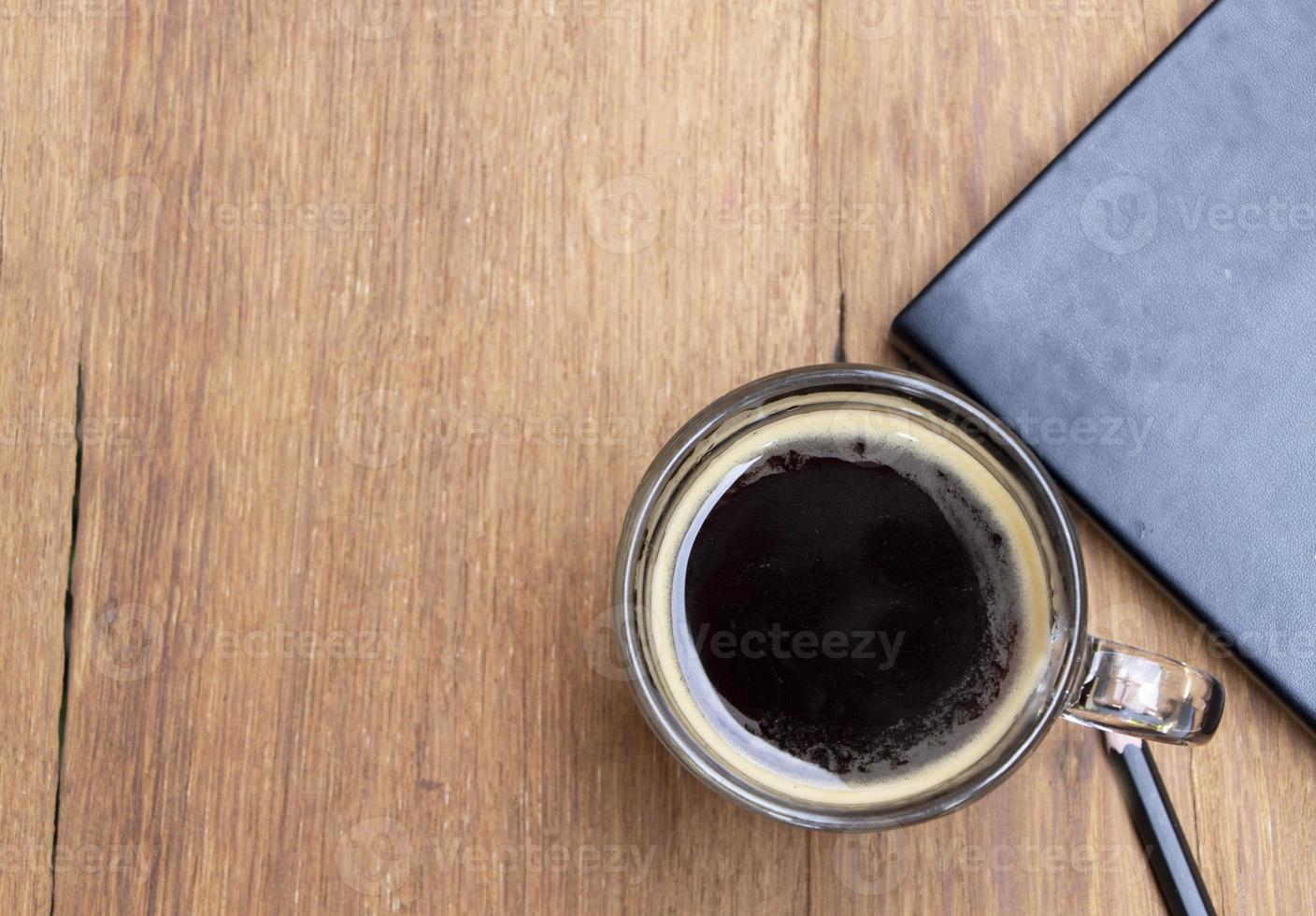 ovanifrån av svart hett americano kaffe i en kopp på ett rustikt träbord med en bok och en penna. koppla av och kaffepaus koncept foto