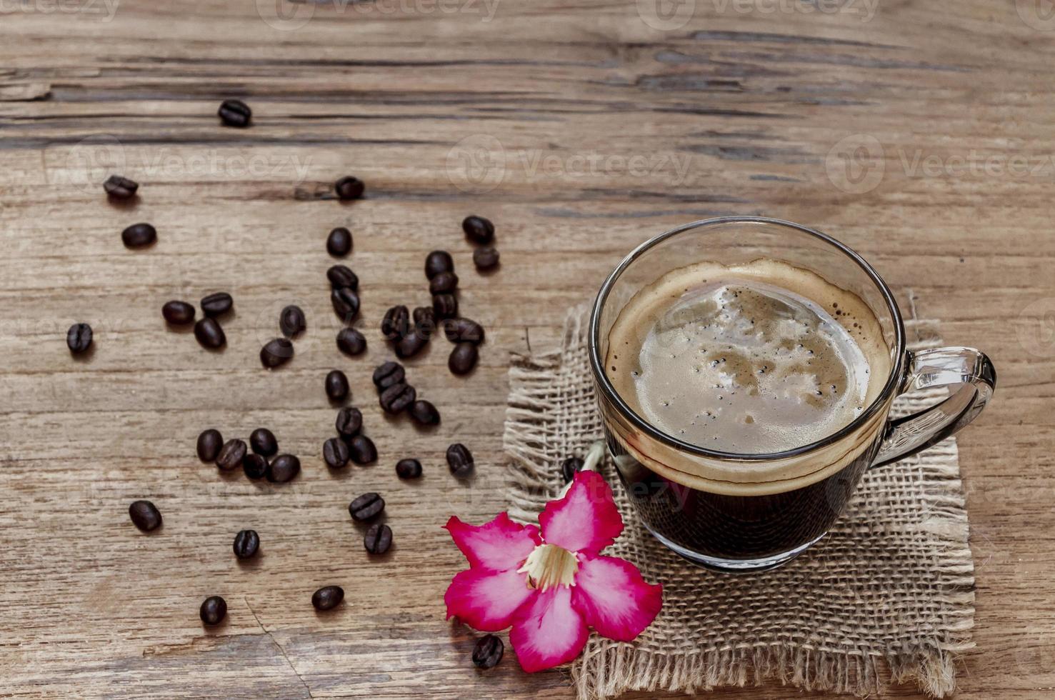 högvinkelvy av en kopp svart americano kaffe på ett rustikt träbord av säckväv med en blomma och en hög med ekologiska bruna kaffebönor. morgon drinkar koncept foto