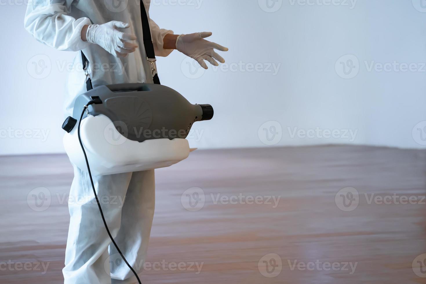 professionell teknisk man i förebyggande kostym sprayar steriliseringslösning med elektrisk spraymaskin på trägolvet och vit bakgrund med studioljus. foto